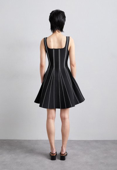 Robe noire avec un corsage ajusté et une jupe évasée, présentant des rangées verticales de clous en argent sur le devant et le dos. Texture lisse, sans manches.