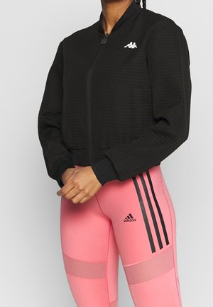 Persona vistiendo una chaqueta negra con cremallera y un logo blanco de Kappa, y leggings rosas de Adidas con rayas negras y paneles de malla.