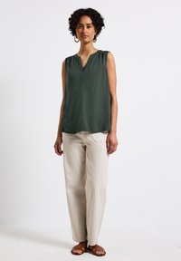 Tmavě zelený bezrukávový top s véčkovým výstřihem, doplněný o světlé béžové kalhoty a hnědé sandály, na jednobarevném bílém pozadí.