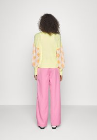 Vrouw met krullend haar die wegkijkt, draagt een lichtgeel truitje met oranje patronen op de mouwen en brede roze broek.