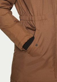 Brown puffer jacket with an adjustable waist, featuring a side pocket with a snap closure and black knit cuff accents. Smooth textured fabric.