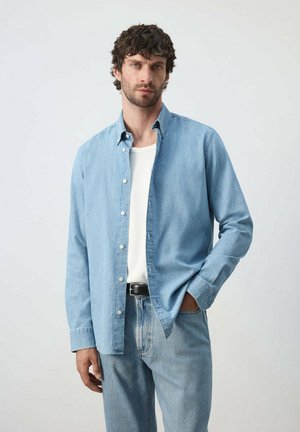Hombre con cabello rizado y oscuro, que lleva una camisa vaquera azul claro sobre una camiseta blanca y vaqueros azules, de pie con una mano en el bolsillo.