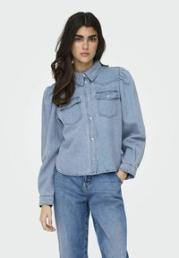 Camicia di denim blu chiaro con colletto, pattina con bottoni, due tasche sul petto e maniche lunghe a sbuffo, abbinata a jeans blu.