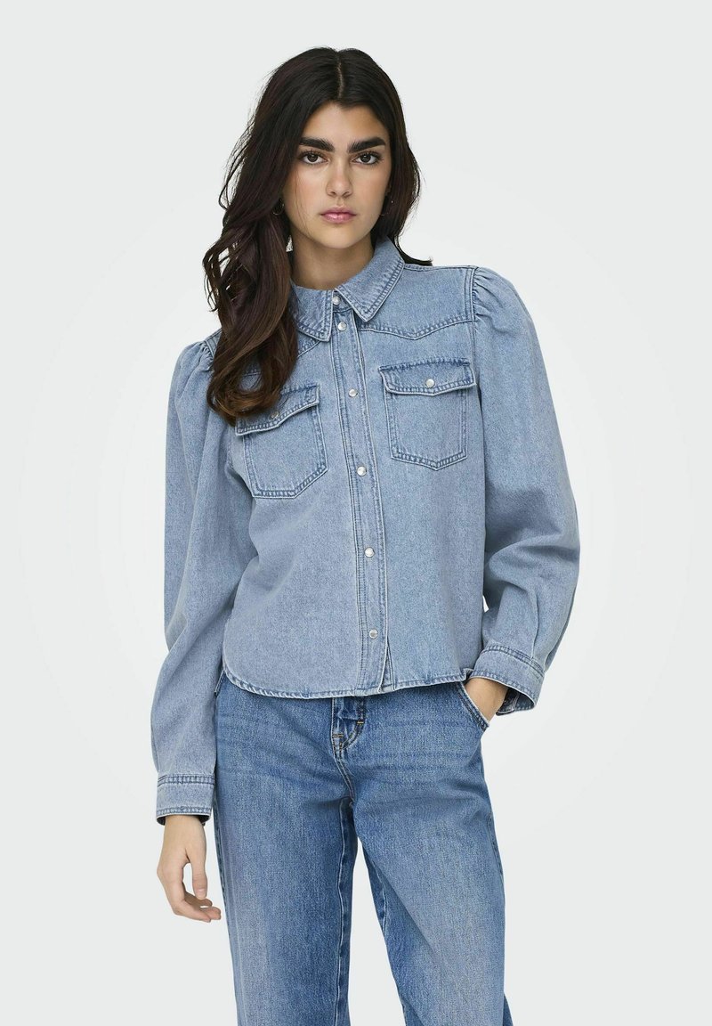 Camicia di denim blu chiaro con colletto, pattina con bottoni, due tasche sul petto e maniche lunghe a sbuffo, abbinata a jeans blu.