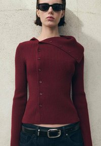 Maglione bordeaux a coste con colletto asimmetrico unico, chiusure con bottoni e maniche lunghe, abbinato a una cintura nera e jeans.