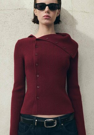Pull en tricot côtelé bordeaux avec un col asymétrique unique, des boutons-pression et des manches longues, associé à une ceinture noire et un jean.