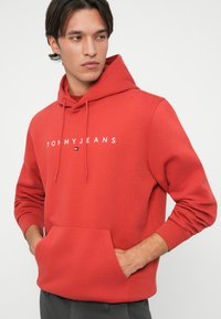 Červená bavlnená mikina s kapucňou, predným vreckom, sťahovacou šnúrkou a bielym textom "TOMMYJEANS". Má ribbované rukávy a uvoľnený strih.