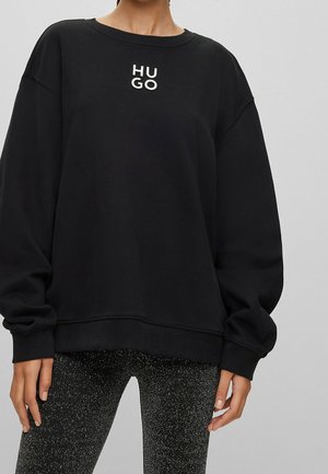 Persoon draagt een zwarte oversized trui met "HUGO" in witte letters op de borst en zwarte gespikkelde legging, staand tegen een grijze achtergrond.