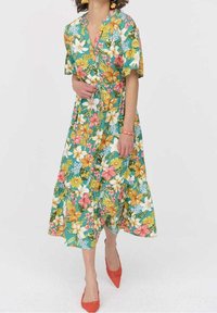 Robe portefeuille florale avec une base turquoise ornée de fleurs multicolores, manches courtes, taille nouée et ourlet à volants. Chaussures rouges décoratives.