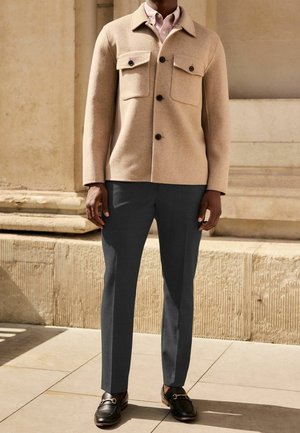 Homme portant une veste en laine beige boutonnée, une chemise rose clair, un pantalon gris foncé et des mocassins en cuir noir, debout dehors près d'un mur en pierre.