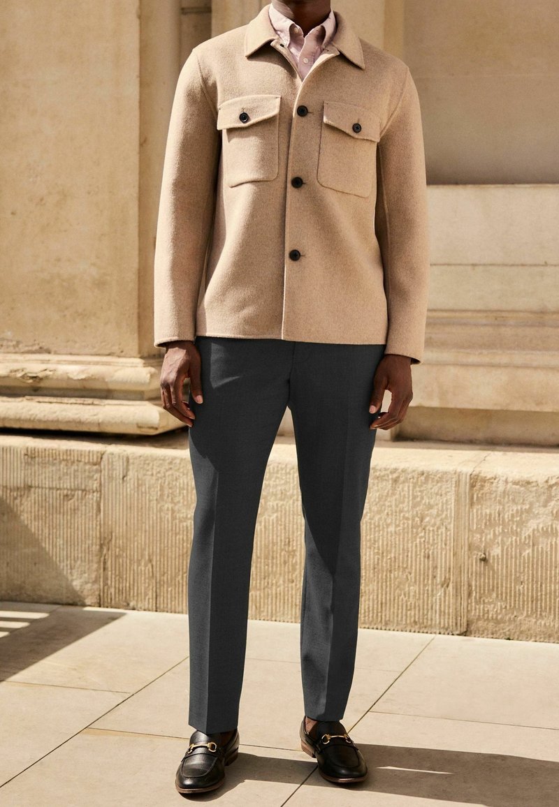 Homme portant une veste en laine beige boutonnée, une chemise rose clair, un pantalon gris foncé et des mocassins en cuir noir, debout dehors près d'un mur en pierre.
