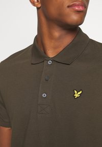 Polo-shirt vert foncé en tissu texturé. Il possède un col, trois boutons et un logo d'oiseau brodé jaune sur le côté gauche de la poitrine.