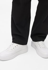 Fehér bőr sportcipők sima felülettel, kerek orral és megemelt fehér talppal, fekete szövetnadrággal párosítva. Fűzős, fűzőlyukakon keresztül.