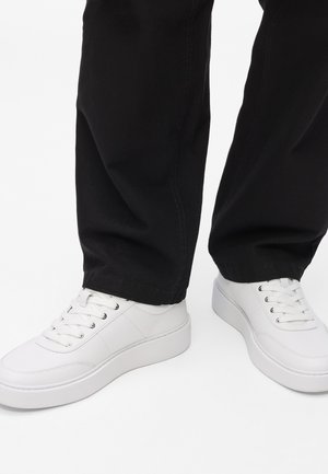 Baltas ādas sporta apavi ar gludu tekstūru, apaļu purngalu un paceltu baltu zoli, kombinēti ar melniem auduma biksēm. Šņores caur acu caurumiem.