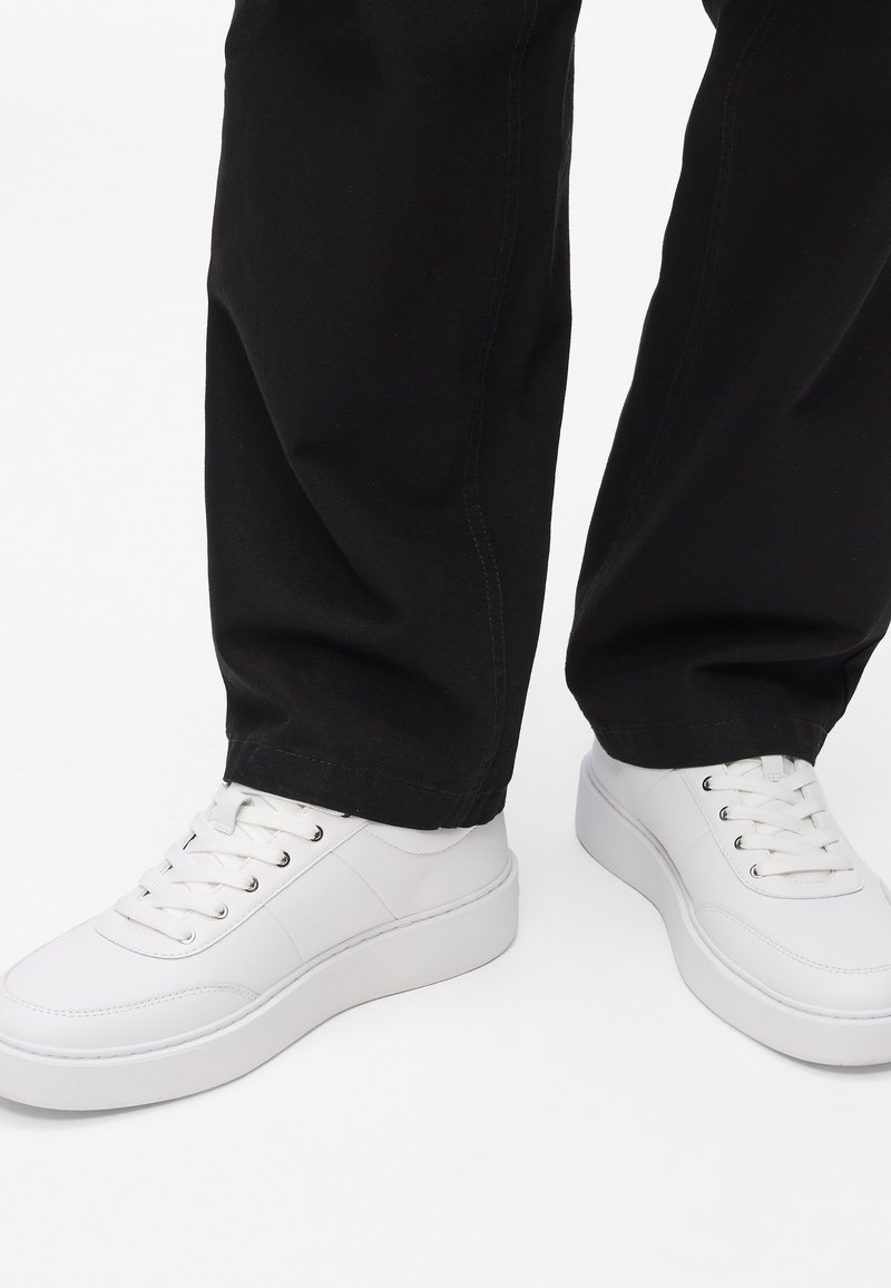 Fehér bőr sportcipők sima felülettel, kerek orral és megemelt fehér talppal, fekete szövetnadrággal párosítva. Fűzős, fűzőlyukakon keresztül.