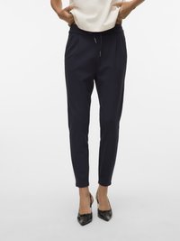 Pantaloni conici bleumarin cu pliuri, talie elastică și detaliu cu șnur; asortați cu pantofi negri cu vârf ascuțit și un top deschis la culoare.