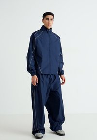 Navy blue athletic jacket and pants set made of lightweight material, featuring contrasting light gray stitching and an elastic waistband.