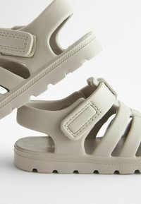 Beige Sandalen mit offenen Seiten, verstellbaren Riemen, flachen Sohlen und strukturierten Gummisohlen mit einem Zickzackmuster für besseren Grip.