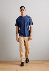La camiseta marina con un pequeño logo, pantalones beige y zapatos negros. El modelo lleva una gorra marina a juego con un borde de color claro.