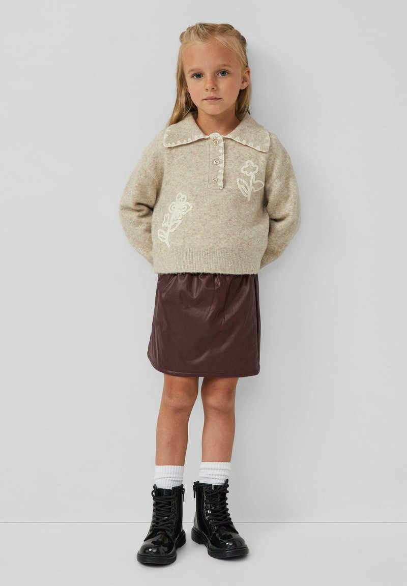 Pull beige avec broderies florales, patte de boutonnage et col festonné, associé à une jupe bordeaux brillante et des bottes noires à lacets.