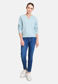 Hellblaues Strickpullover mit V-Ausschnitt, der ein strukturiertes Rautenmuster aufweist, kombiniert mit dunkelblauen Jeans und weißen Sneakers.