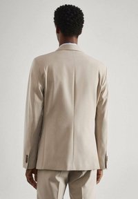 Beige colbert met een getailleerde pasvorm en een enkele split aan de achterkant. Voorzien van knoopsluiting bij de mouwen en een gladde stofstructuur.