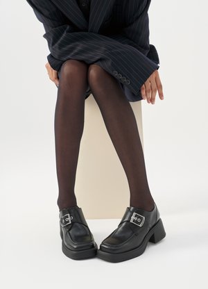 Person trägt schwarzen Nadelstreifenblazer, schwarze transparente Strumpfhosen und schwarze Leder-Pumps mit silbernen Schnallen, sitzt auf einem hellen Block.