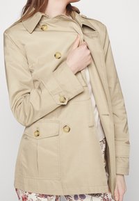 Beige trenčkot s dvojitým zapínáním, zlatými knoflíky a klopovými kapsami. Hladká textura, klasický límec, mírně zúžený pas.