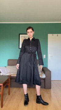 Vestido preto com botões, mangas compridas, cinto na cintura e saia plissada. Usado com botas pretas até ao tornozelo com fivelas decorativas.