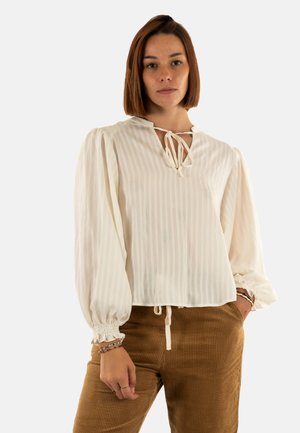 Mujer con cabello castaño vistiendo una blusa blanca holgada con lazos y mangas abullonadas, y pantalones de pana marrón, de pie contra un fondo liso.