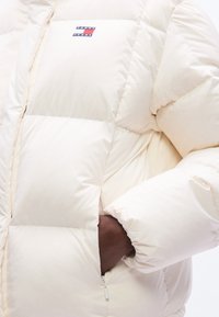 Φουσκωτό μπεζ μπουφάν με σχεδιασμό σε ραμμένη μοτίβα, με επωμίδα λογότυπου και τσεπές στο πλάι με φερμουάρ. Μαλακή υφή, φαρδιά γραμμή.