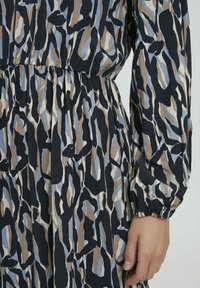 Robe à motifs noir, bleu et beige avec des manches longues. Le tissu est léger avec une taille élastique. Présente une gamme de formes abstraites.