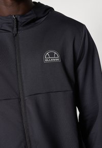 Gros plan sur une personne portant un sweat à capuche zippé noir avec un logo Ellesse blanc sur la poitrine gauche.