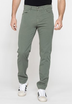 Uomo che indossa pantaloni slim verde oliva e sneakers bianche con strisce nere, in piedi su uno sfondo chiaro e uniforme.