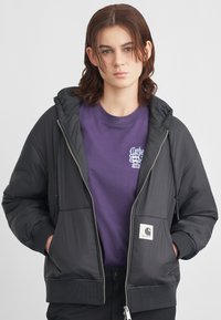 Schwarze gepolsterte Jacke mit Kapuze, gerippten Bündchen und Seitentaschen. Getragen über einem lila T-Shirt mit einem weißen Grafiklogo.