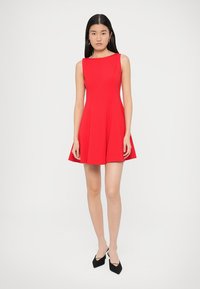 Rotes ärmelloses Kleid mit tailliertem Oberteil und ausgestelltem Rock. Hergestellt aus glattem Stoff, mit klassischer A-Linien-Form. Kombiniert mit schwarzen High Heels.