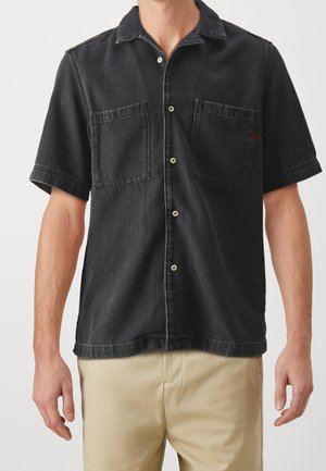 Mężczyzna ubrany w czarną koszulę na krótki rękaw z dwoma kieszeniami na piersi i beżowe spodnie na jednolitym tle.