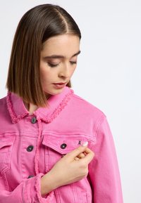 Chaqueta de mezclilla rosa con cuello y puños deshilachados, presenta botones negros y detalles bordados. El modelo toca el bolsillo del pecho.