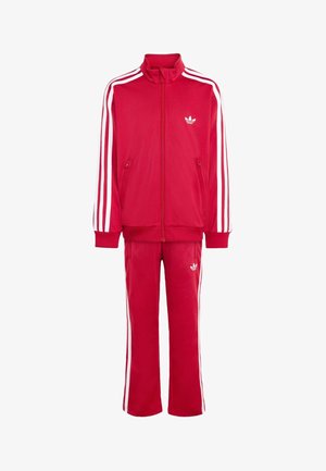 Czerwony dres Adidas z białymi trzema paskami na rękawach i spodniach, bluza na zamek i logo na piersi oraz udzie.