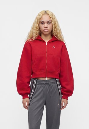 Jonge persoon met lang krullend blond haar, die een rode cropped hoodie en grijze sportbroek met witte strepen en logo’s draagt.