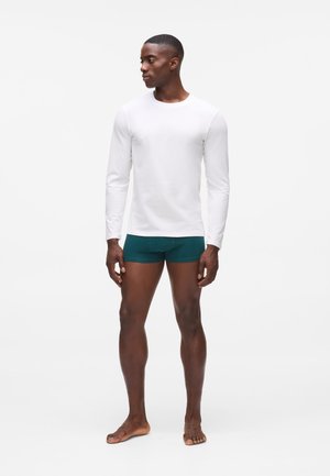 Chemise blanche à manches longues associée à un short en boxeur vert foncé. Modèle debout pieds nus, mettant en valeur un design ajusté et une texture de tissu lisse.