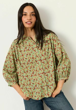 Frau mit langen dunklen Haaren, die eine grüne Bluse mit rotem Blumenmuster und blaue Jeans trägt, steht mit den Händen in den Taschen vor einer weißen Wand.