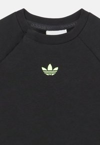 Crna dukserica s rebrastim okruglim izrezom i malim, reflektirajućim zelenim Adidas trefoil logotipom na prsima. Mekani materijal.
