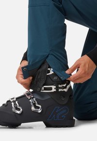 Scarponi da sci neri con fibbie argentate e dettagli blu, abbinati a pantaloni color teal. L'individuo sta regolando l'orlo del pantalone attorno allo scarpone.
