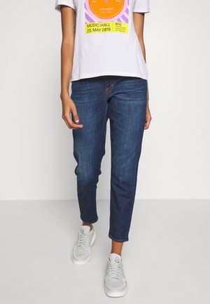 Tmavě modré džíny s tapered střihem, s oranžovým prošíváním, v kombinaci se světle šedým tričkem s potiskem a bílými teniskami.