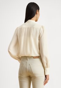 Krém színű blúz strukturált gallérral, összegyűrt hátoldalú résszel és hosszú, puffos ujjakkal, világos bézs színű, testhezálló farmerral párosítva.