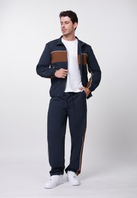 Νεαρός άνδρας που διορθώνει μπλε ναυτικό και καφέ τζάκετ με φερμουάρ, φοράει παντελόνια φόρμας που ταιριάζουν, λευκό μπλουζάκι και λευκά αθλητικά παπούτσια, στεκόμενος σε λευκό φόντο.