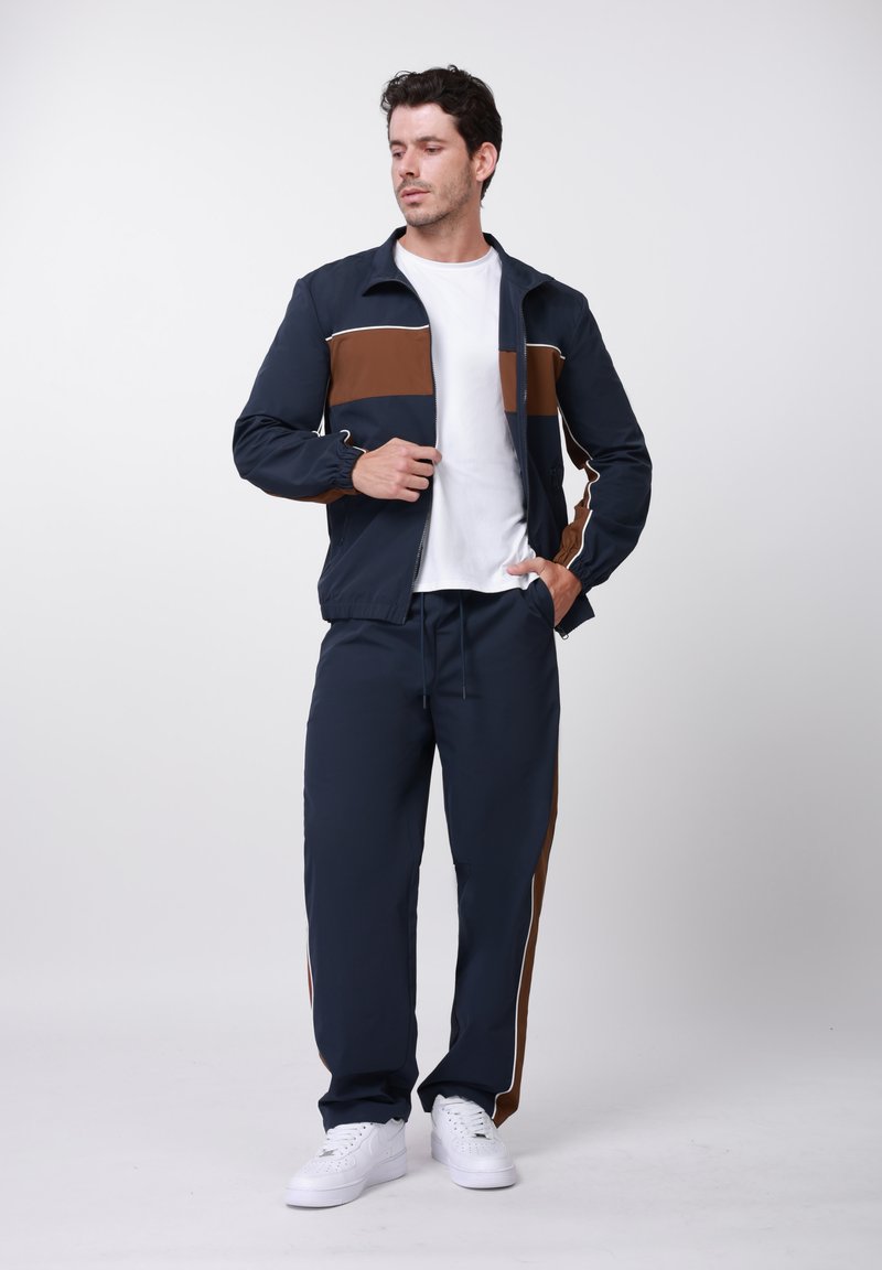 Νεαρός άνδρας που διορθώνει μπλε ναυτικό και καφέ τζάκετ με φερμουάρ, φοράει παντελόνια φόρμας που ταιριάζουν, λευκό μπλουζάκι και λευκά αθλητικά παπούτσια, στεκόμενος σε λευκό φόντο.