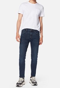 Mężczyzna w białej koszulce z krótkim rękawem z kieszonką, ciemnoniebieskich slim fit dżinsach i białych oraz szarych sneakersach, stojący na białym tle.