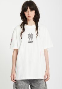 Camiseta de algodón blanca con mangas cortas, que presenta un diseño gráfico negro en el pecho y detalles coloridos en la manga izquierda.